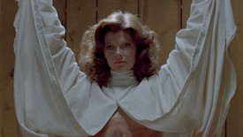 A woman stares forward and stands with her arms raised and draped in a white cloak.