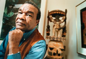Dr. David Driskell, 2002, head resting on hand in a blue shirt with art in the background.