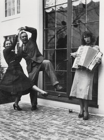Black-and-white photograph of Alexander Calder and Margaret French dancing on a cobblestone street while Louisa Calder plays the accordion in front of a large window outside of James Thrall Soby’s house, Farmington, Connecticut, 1936