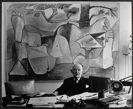 A black-and-white portrait of Daniel-Henry Kahnweiler seated at a desk in front of a painting by Pablo Picasso.